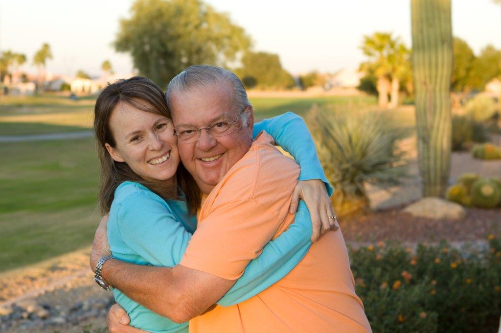 Senior father hugging adult daughter - enjoying senior living in Arizona with grass, cactus and desert landscaping behind them
